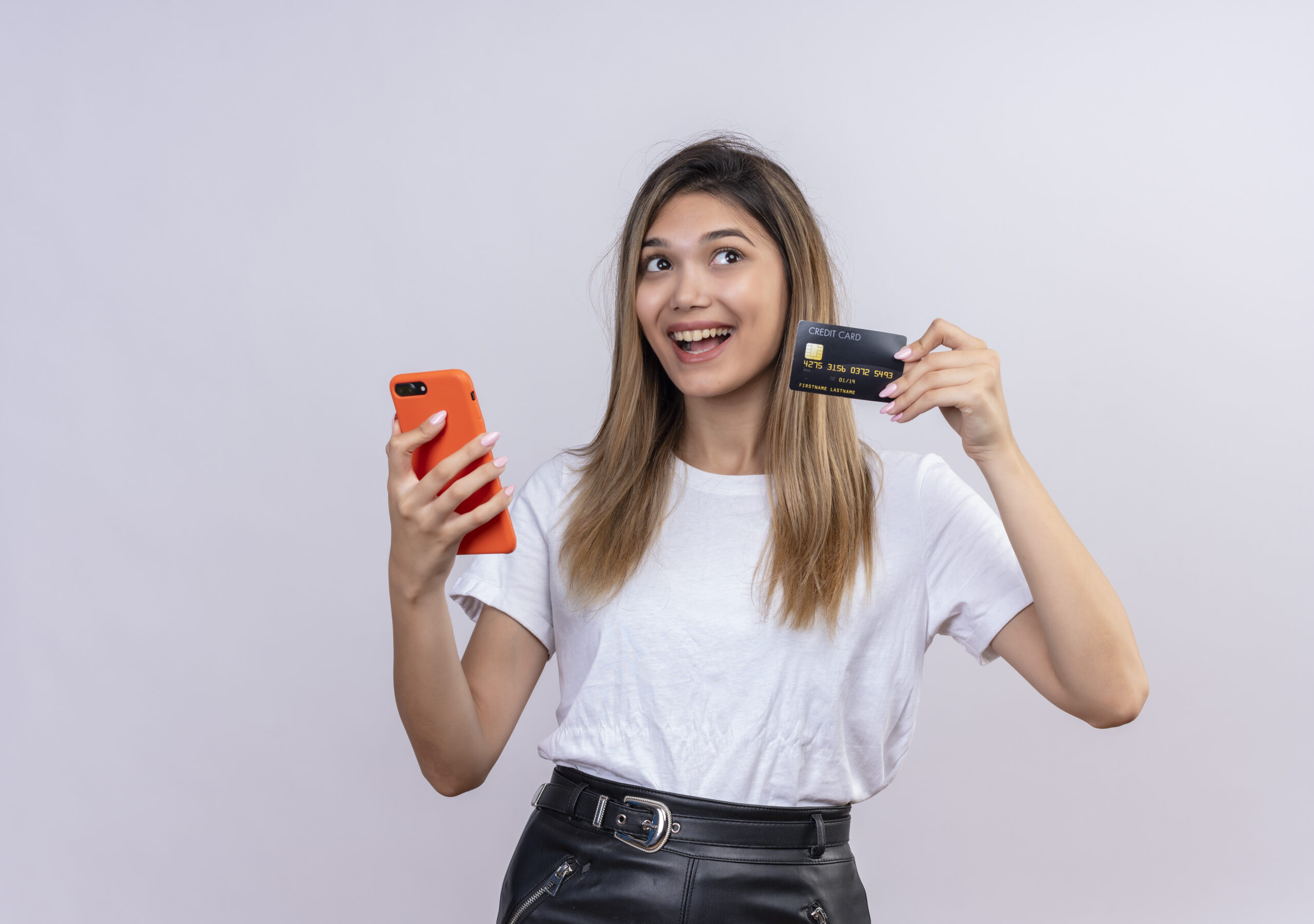 a cheerful young woman in white t-shirt showing credit card while holding mobile phone on a white background